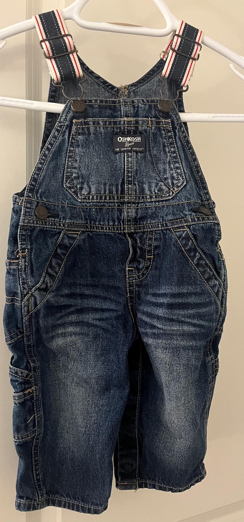 Puede incluir: Pantal&oacute;n vaquero azul con tirantes ajustables con rayas rojas y blancas. El pantal&oacute;n tiene un parche en la parte delantera que dice "OSHKOSH B'Gosh THE GENUINE ARTICLE".