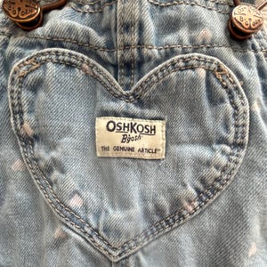 K&ouml;nnte beinhalten: Nahaufnahme einer hellblauen Jeans-Latzhose mit einer herzf&ouml;rmigen Tasche. Die Latzhose tr&auml;gt das Label "OshKosh B'gosh" und bronzefarbene Beschl&auml;ge. Der Jeansstoff hat ein verwaschenes Aussehen mit wei&szlig;en Sprenkeln.