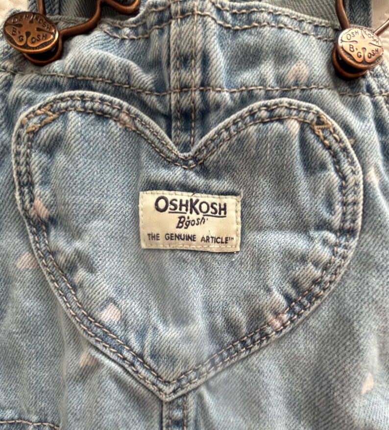 K&ouml;nnte beinhalten: Nahaufnahme einer hellblauen Jeans-Latzhose mit einer herzf&ouml;rmigen Tasche. Die Latzhose tr&auml;gt das Label "OshKosh B'gosh" und bronzefarbene Beschl&auml;ge. Der Jeansstoff hat ein verwaschenes Aussehen mit wei&szlig;en Sprenkeln.