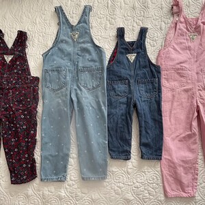 K&ouml;nnte beinhalten: Vier Kinder-Latzhosen in verschiedenen Farben und Mustern. Ein Paar ist dunkelrot mit floralem Muster, ein anderes hellblau mit wei&szlig;en Punkten, ein drittes dunkelblauer Denim und das letzte rosa.