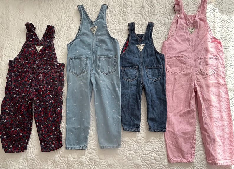 K&ouml;nnte beinhalten: Vier Kinder-Latzhosen in verschiedenen Farben und Mustern. Ein Paar ist dunkelrot mit floralem Muster, ein anderes hellblau mit wei&szlig;en Punkten, ein drittes dunkelblauer Denim und das letzte rosa.