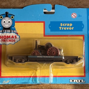 May include: A Thomas & Friends toy featuring Scrap Trevor, a dark brown steam roller with red wheels, on a flatbed. The packaging is blue and yellow, with the Thomas & Friends logo and the text "SAVED FROM SCRAP".