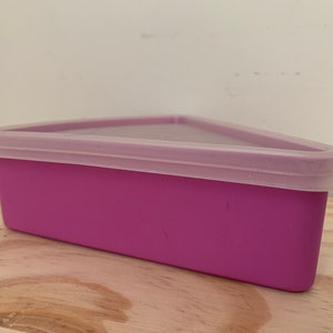 May include: A triangular, magenta-colored food storage container with a translucent, light purple lid. The container is made of plastic and sits on a wooden surface. The lid fits snugly on top of the container.