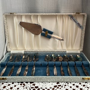 Könnte beinhalten: Ein silberner Kuchenspachtel und 10 silberne Teelöffel sind in einer mit blauem Samt ausgekleideten Schachtel mit weißem Blumenmuster ausgestellt. Die Schachtel ist geöffnet und der Spachtel und die Löffel sind in ihren dafür vorgesehenen Fächern angeordnet. Die Schachtel ist mit "Yoeman Plate" beschriftet.