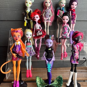 May include: A selection of Monster High dolls, each with distinctive characteristics and attire. The dolls display diverse skin tones, hair colours, and clothing styles, including dresses, skirts, and tops. Some dolls have animal-inspired features, such as cat ears and tails.