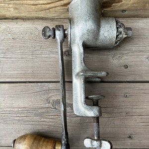 Könnte beinhalten: Ein antiker, silberfarbener manueller Fleischwolf mit einem Holzgriff. Der Fleischwolf hat eine Klemme, um ihn an einer Oberfläche zu befestigen, und eine Kurbel, um den Mahlmechanismus zu drehen.