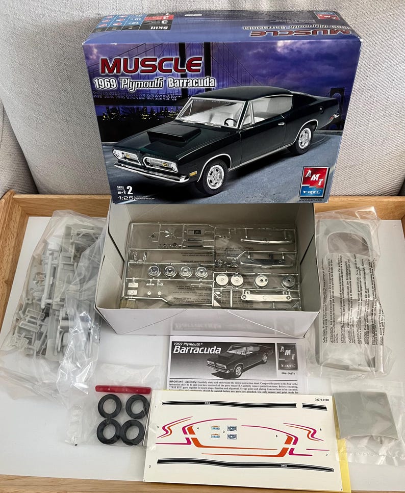 Puede incluir: Un kit de maqueta de un Plymouth Barracuda 1969 en verde. La caja muestra el coche en una foto en blanco y negro. El kit incluye piezas, instrucciones y calcoman&iacute;as. La caja est&aacute; etiquetada como "Muscle".