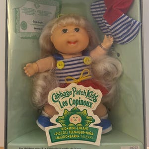 Pode incluir: Uma boneca Cabbage Patch Kids de cabelo loiro, vestindo um top às riscas azuis e brancas com botões amarelos e uma saia vermelha. A boneca inclui um chapéu às riscas vermelhas e brancas a condizer, numa caixa com o texto Cabbage Patch Kids Les Copinoux.
