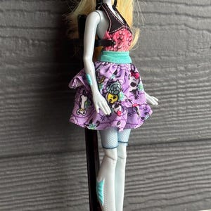 May include: A Monster High doll with pale skin, blonde hair, and pink lips. She wears a black and pink top, a purple skirt with skull and cartoon graphics, and bright pink shoes. The doll is standing on a black stand.