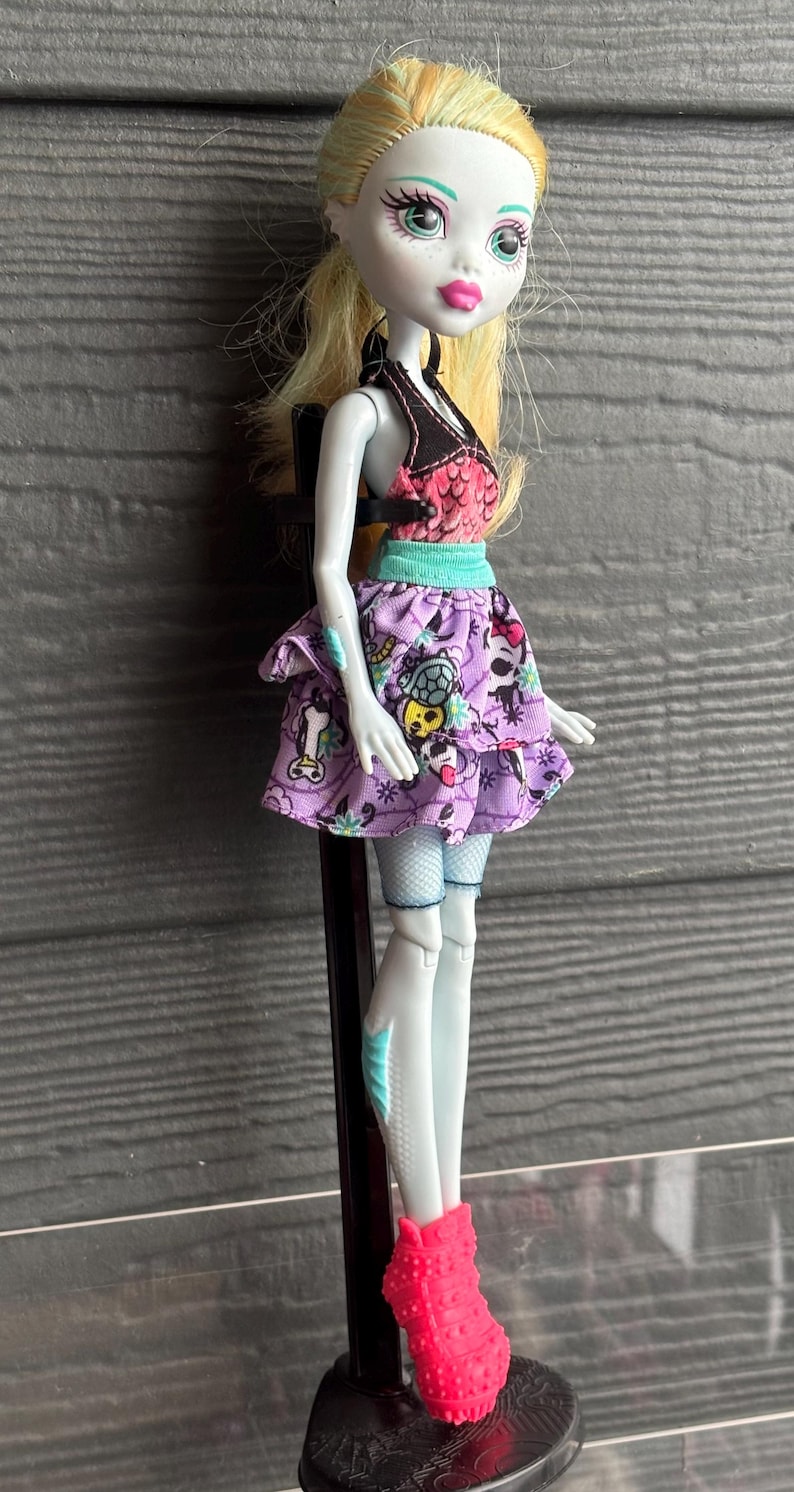 May include: A Monster High doll with pale skin, blonde hair, and pink lips. She wears a black and pink top, a purple skirt with skull and cartoon graphics, and bright pink shoes. The doll is standing on a black stand.