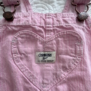 K&ouml;nnte beinhalten: Rosa und wei&szlig; gestreifte Latzhose mit herzf&ouml;rmiger Tasche. Die Latzhose hat verstellbare Tr&auml;ger mit bronzefarbener Hardware. Die Tasche hat ein wei&szlig;es Etikett mit dem Markennamen "OshKosh B'gosh" und dem Text "The Genuine Article."