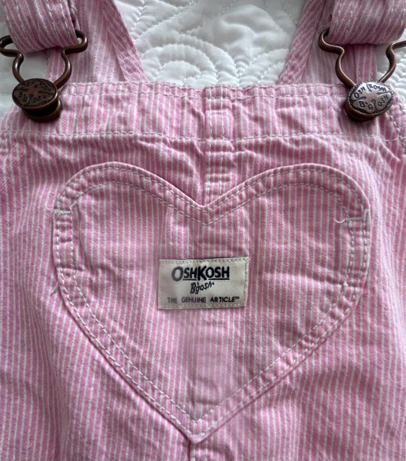 K&ouml;nnte beinhalten: Rosa und wei&szlig; gestreifte Latzhose mit herzf&ouml;rmiger Tasche. Die Latzhose hat verstellbare Tr&auml;ger mit bronzefarbener Hardware. Die Tasche hat ein wei&szlig;es Etikett mit dem Markennamen "OshKosh B'gosh" und dem Text "The Genuine Article."