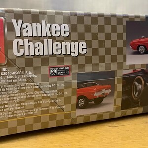 Op de afbeelding: Een modelbouwdoos voor een rode Dodge Challenger Yankee Challenge uit 1970. De doos bevat afbeeldingen van de auto, waaronder een close-up van de motor en het interieur. De doos bevat ook de tekst "Yankee Challenge" en "Dodge Grab Life by the Horns".