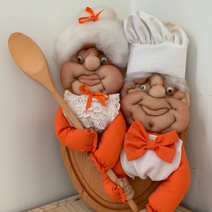 May include: A pair of handmade fabric dolls depicting a chef and a baker. The dolls are wearing white hats and orange aprons. The baker is holding a wooden spoon. The dolls are mounted on a wooden plaque.
