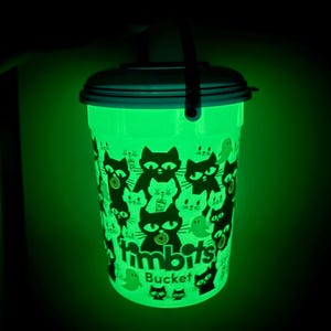 Puede incluir: Cubo de plástico verde que brilla en la oscuridad con un diseño de gato negro y fantasma. El cubo tiene un asa y una tapa. El texto "Timbits Bucket" está impreso en el lateral. El cubo brilla intensamente en la oscuridad.