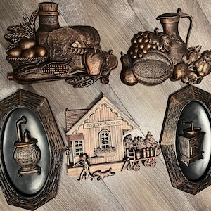 May include: Set of six vintage wall plaques featuring a farmhouse theme. The plaques are made of a textured, metallic material and feature a variety of scenes, including a farmhouse, a basket of fruit, a pitcher, a stove, and a mill. The plaques are painted in shades of brown and gold.