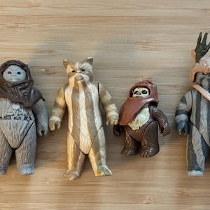 Puede incluir: Una colección de figuras de acción de Star Wars, incluyendo a Chewbacca, Ewoks y un Stormtrooper. Las figuras están en varias poses y presentan diseños detallados. La paleta de colores incluye tonos de marrón, gris y blanco, con algunos personajes con patrones de rayas.