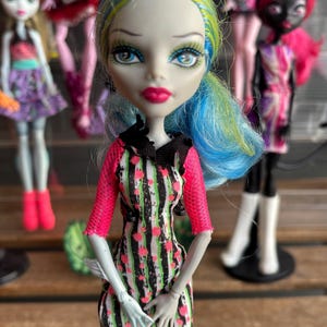 May include: A doll with blue and green hair, wearing a black, green, and pink striped dress with pink sleeves. The doll has pale skin, blue eyes, and pink lipstick. Other dolls are visible in the background.