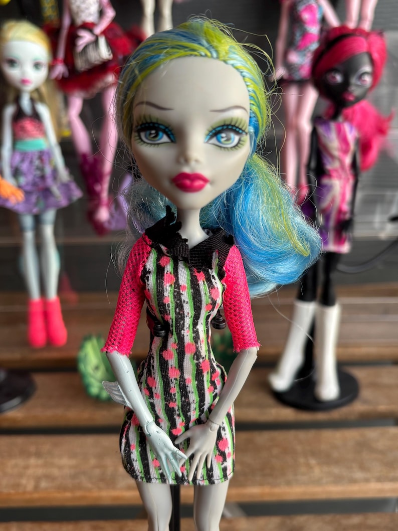 May include: A doll with blue and green hair, wearing a black, green, and pink striped dress with pink sleeves. The doll has pale skin, blue eyes, and pink lipstick. Other dolls are visible in the background.