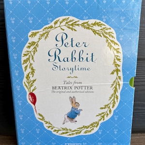 Puede incluir: Un estuche azul de libros "Peter Rabbit Storytime". La portada presenta un marco blanco con el texto "Peter Rabbit Storytime" y "Tales from Beatrix Potter". También se muestra un conejo de dibujos animados con una chaqueta azul.