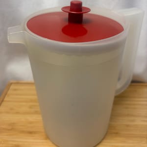 May include: A clear plastic jug with a red lid and handle. The jug has a spout for easy pouring. The lid has a small red knob on top. The jug is sitting on a wooden surface.