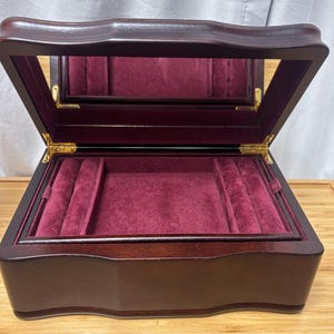 May include: A wooden jewelry box with a hinged lid and a built-in mirror. The interior is lined with a deep red velvet material. The box is open, revealing compartments for storing jewelry. The box is made of dark brown wood.