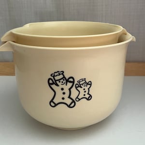 Peut inclure: Deux saladiers crème, l'un niché dans l'autre. Chaque bol a une poignée et un dessin noir d'un bonhomme en pain d'épices. Les bols sont en matériau durable, parfaits pour la pâtisserie et la préparation des aliments.