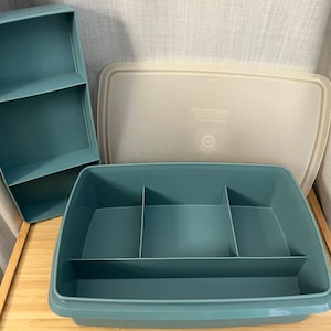 Può includere: Un set di contenitori in plastica color verde acqua con coperchio e un organizer separato a due ripiani. Il contenitore ha divisori interni. Il coperchio è bianco sporco. Il set è progettato per l'organizzazione e lo stoccaggio.