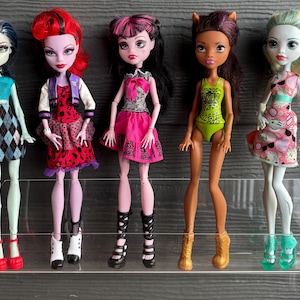 May include: Five Monster High dolls are displayed. The dolls have unique features, including different skin tones, hair colours, and outfits. One doll wears a blue and black diamond-patterned dress, while another has a red and black spotted dress. The dolls are on a clear shelf.