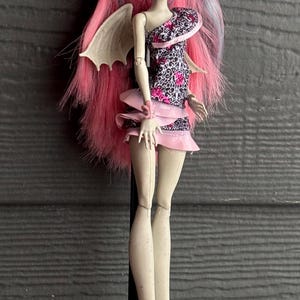 May include: A doll with pink and blue hair, wearing a black and pink patterned dress and a pink skirt. The doll has white wings and white boots with pink heels. The doll is a collectible item.