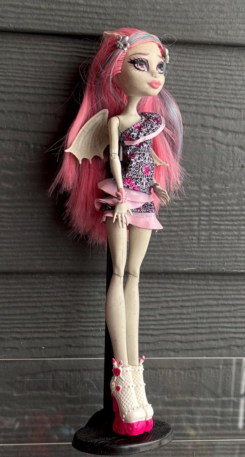 May include: A doll with pink and blue hair, wearing a black and pink patterned dress and a pink skirt. The doll has white wings and white boots with pink heels. The doll is a collectible item.