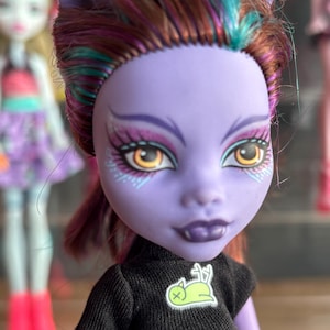 May include: A doll with a purple face, dark hair with teal and pink highlights, and yellow eyes. The doll wears a black shirt with a green and white graphic. The doll has pointed ears and fangs. Other dolls are in the background.