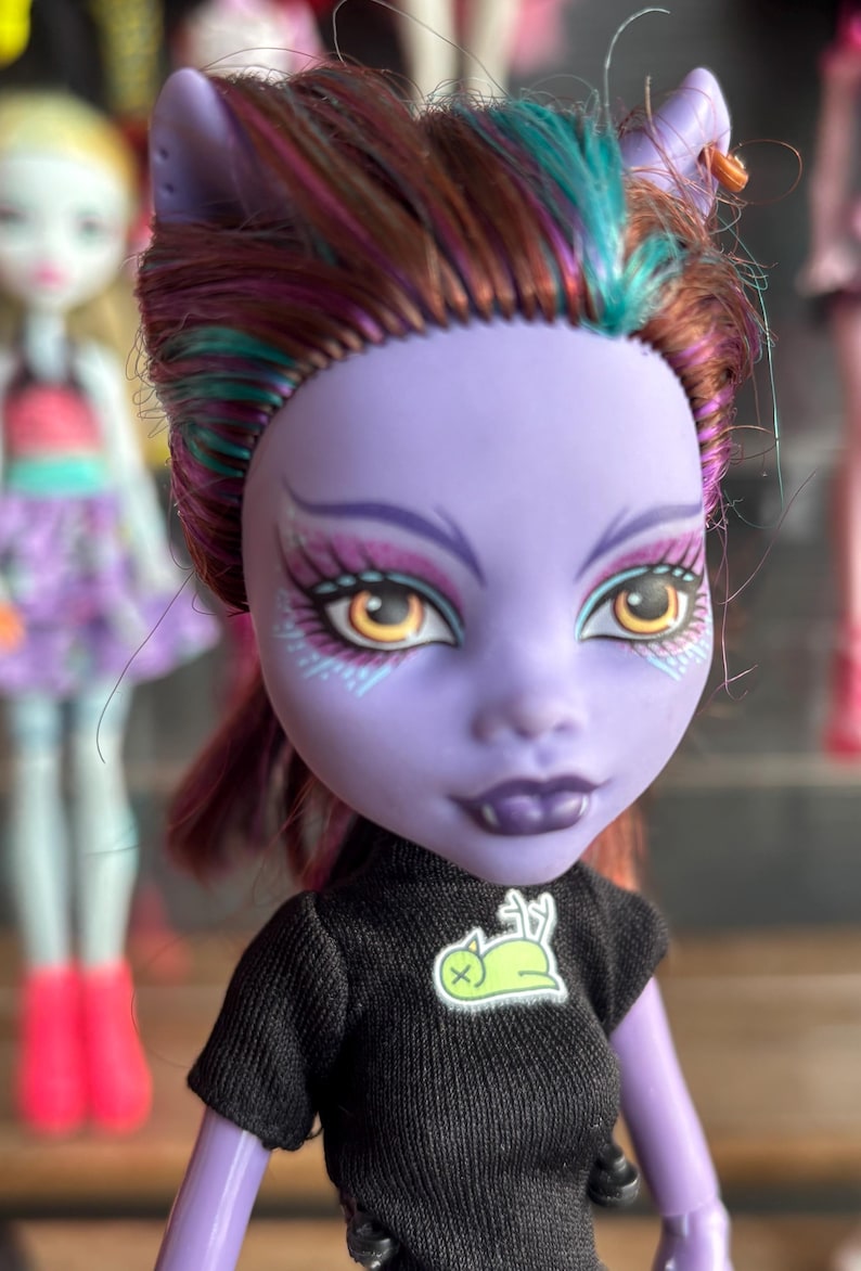 May include: A doll with a purple face, dark hair with teal and pink highlights, and yellow eyes. The doll wears a black shirt with a green and white graphic. The doll has pointed ears and fangs. Other dolls are in the background.