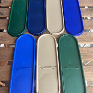 May include: An overhead shot of eight plastic Tupperware lids in various colors: green, navy blue, beige, and royal blue. The lids are oval-shaped and have a hinged design. The lids are arranged on a wooden surface.