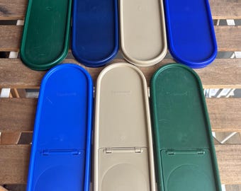 May include: An overhead view of eight plastic Tupperware lids in green, navy blue, beige, and royal blue. The oval-shaped lids have a hinged design. The lids are arranged on a wooden surface.