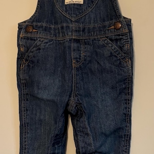 K&ouml;nnte beinhalten: Dunkelblaue Jeans-Latzhose mit herzf&ouml;rmigem Latz und dem Logo "OshKosh B'gosh". Die Latzhose hat verstellbare Tr&auml;ger, Taschen und Knopfverschl&uuml;sse. Das Kleidungsst&uuml;ck h&auml;ngt an einem wei&szlig;en Kleiderb&uuml;gel.