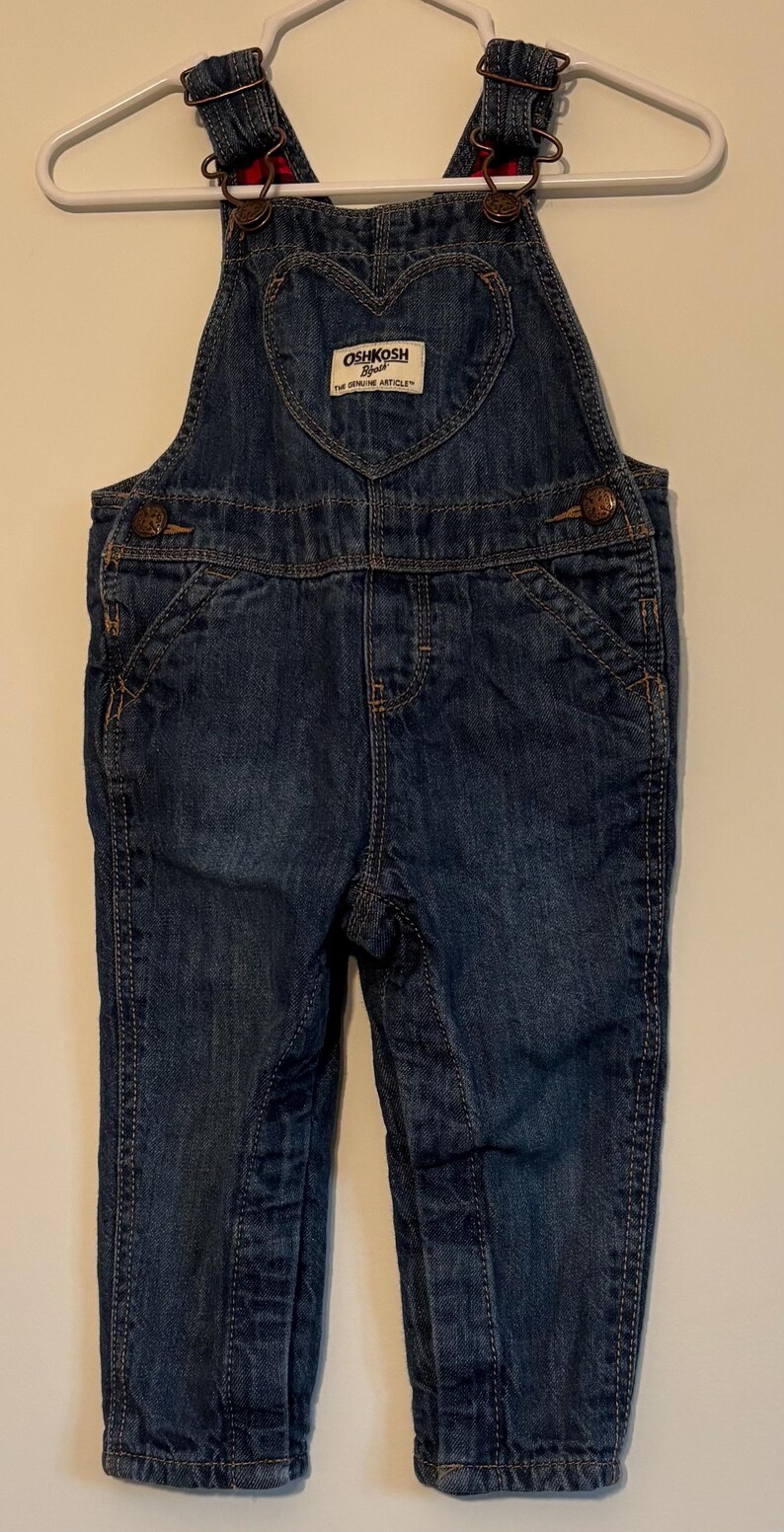 K&ouml;nnte beinhalten: Dunkelblaue Jeans-Latzhose mit herzf&ouml;rmigem Latz und dem Logo "OshKosh B'gosh". Die Latzhose hat verstellbare Tr&auml;ger, Taschen und Knopfverschl&uuml;sse. Das Kleidungsst&uuml;ck h&auml;ngt an einem wei&szlig;en Kleiderb&uuml;gel.