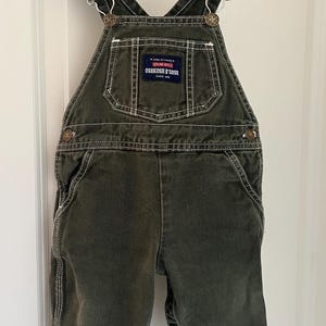 Puede incluir: Pantal&oacute;n vaquero verde con costuras blancas y un parche de logotipo marr&oacute;n OshKosh B'Gosh. El pantal&oacute;n tiene tirantes ajustables con hebillas met&aacute;licas.