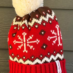 May include: Red knit beanie with a white pom-pom. The hat features a pattern of white snowflakes, brown and white geometric shapes. The brim is solid red, and a white tag with the text "Tim Hortons" is attached.