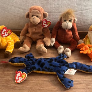 May include: Five plush toys, including a yellow giraffe, a brown monkey, an orange monkey, a green frog, and a blue and yellow lizard. Each toy has a red and white tag attached.