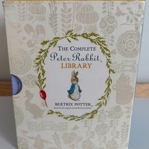 Peut inclure: Un coffret de "The Complete Peter Rabbit Library" de Beatrix Potter. La boîte crème présente un motif floral et une illustration de Pierre Lapin en veste bleue. Le titre et le nom de l'auteur sont bien en évidence.