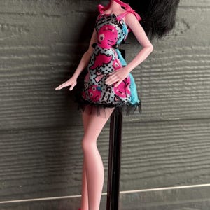 May include: A doll with black hair in a high ponytail, wearing a pink and blue patterned dress with a black lace trim. The doll has pink skin, red lips, and heart-shaped cheek markings. She is wearing pink high-heeled shoes.