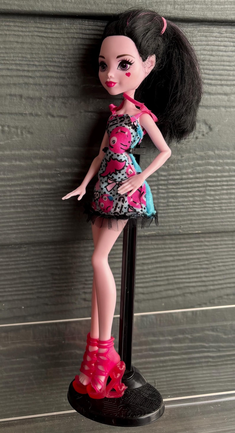 May include: A doll with black hair in a high ponytail, wearing a pink and blue patterned dress with a black lace trim. The doll has pink skin, red lips, and heart-shaped cheek markings. She is wearing pink high-heeled shoes.