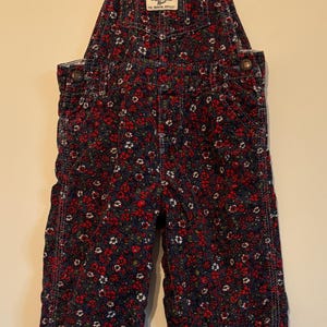 K&ouml;nnte beinhalten: Marineblaue Cord-Latzhose mit rotem und wei&szlig;em Blumenmuster. Die Latzhose hat verstellbare Tr&auml;ger und das Markenetikett "OshKosh B'gosh". Das Kleidungsst&uuml;ck h&auml;ngt an einem wei&szlig;en Kleiderb&uuml;gel, um sein Design und seine Details zu zeigen.