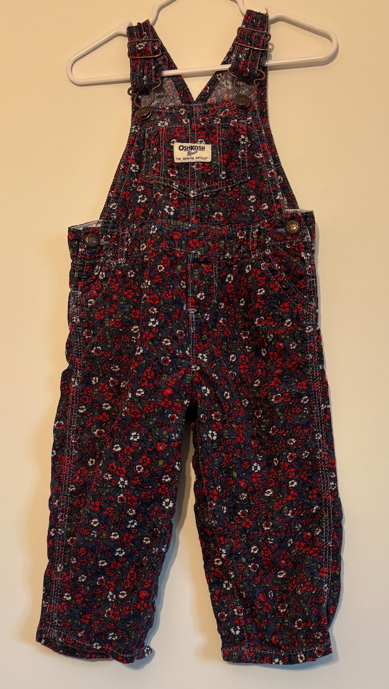 K&ouml;nnte beinhalten: Marineblaue Cord-Latzhose mit rotem und wei&szlig;em Blumenmuster. Die Latzhose hat verstellbare Tr&auml;ger und das Markenetikett "OshKosh B'gosh". Das Kleidungsst&uuml;ck h&auml;ngt an einem wei&szlig;en Kleiderb&uuml;gel, um sein Design und seine Details zu zeigen.