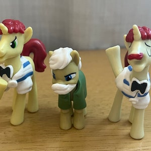 May include: Three toy horses wearing red, white and blue sailor outfits with bow ties. The horses are yellow with white manes and tails. One horse has a white beard.