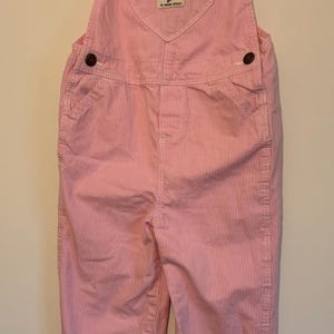 K&ouml;nnte beinhalten: Rosa-wei&szlig; gestreifte OshKosh B'gosh Latzhose mit verstellbaren Tr&auml;gern und braunen Metallbeschl&auml;gen. Das L&auml;tzchen hat ein herzf&ouml;rmiges Design und ein kleines Etikett. Die Latzhose h&auml;ngt an einem wei&szlig;en Kleiderb&uuml;gel.
