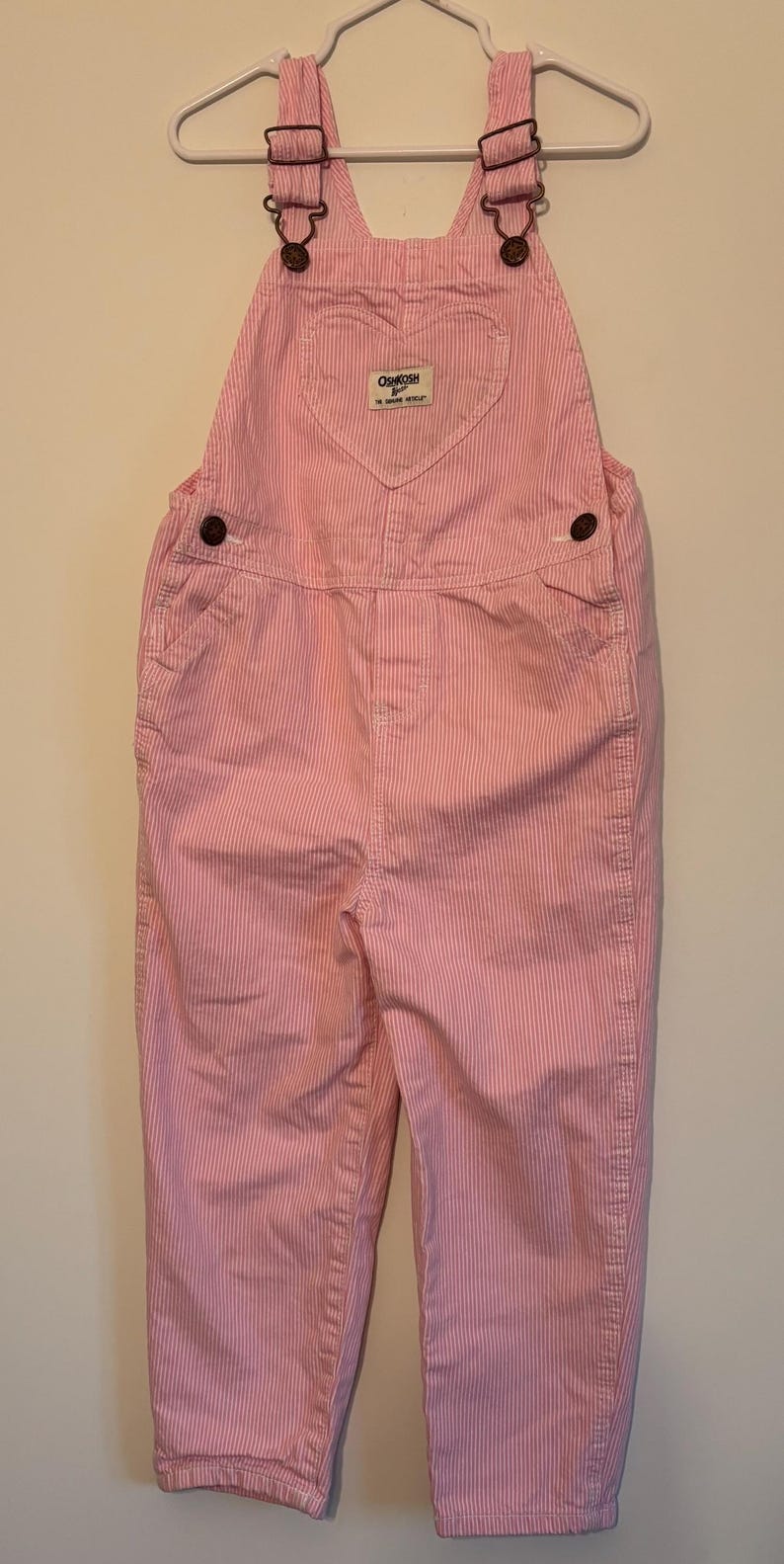 K&ouml;nnte beinhalten: Rosa-wei&szlig; gestreifte OshKosh B'gosh Latzhose mit verstellbaren Tr&auml;gern und braunen Metallbeschl&auml;gen. Das L&auml;tzchen hat ein herzf&ouml;rmiges Design und ein kleines Etikett. Die Latzhose h&auml;ngt an einem wei&szlig;en Kleiderb&uuml;gel.