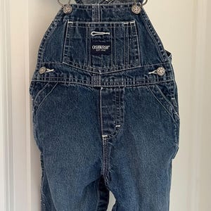 Puede incluir: Pantal&oacute;n vaquero azul para beb&eacute; o ni&ntilde;o peque&ntilde;o con tirantes ajustables y herrajes plateados. El pantal&oacute;n tiene el logotipo de OshKosh B'Gosh en la parte delantera con el texto "EST. 1895".