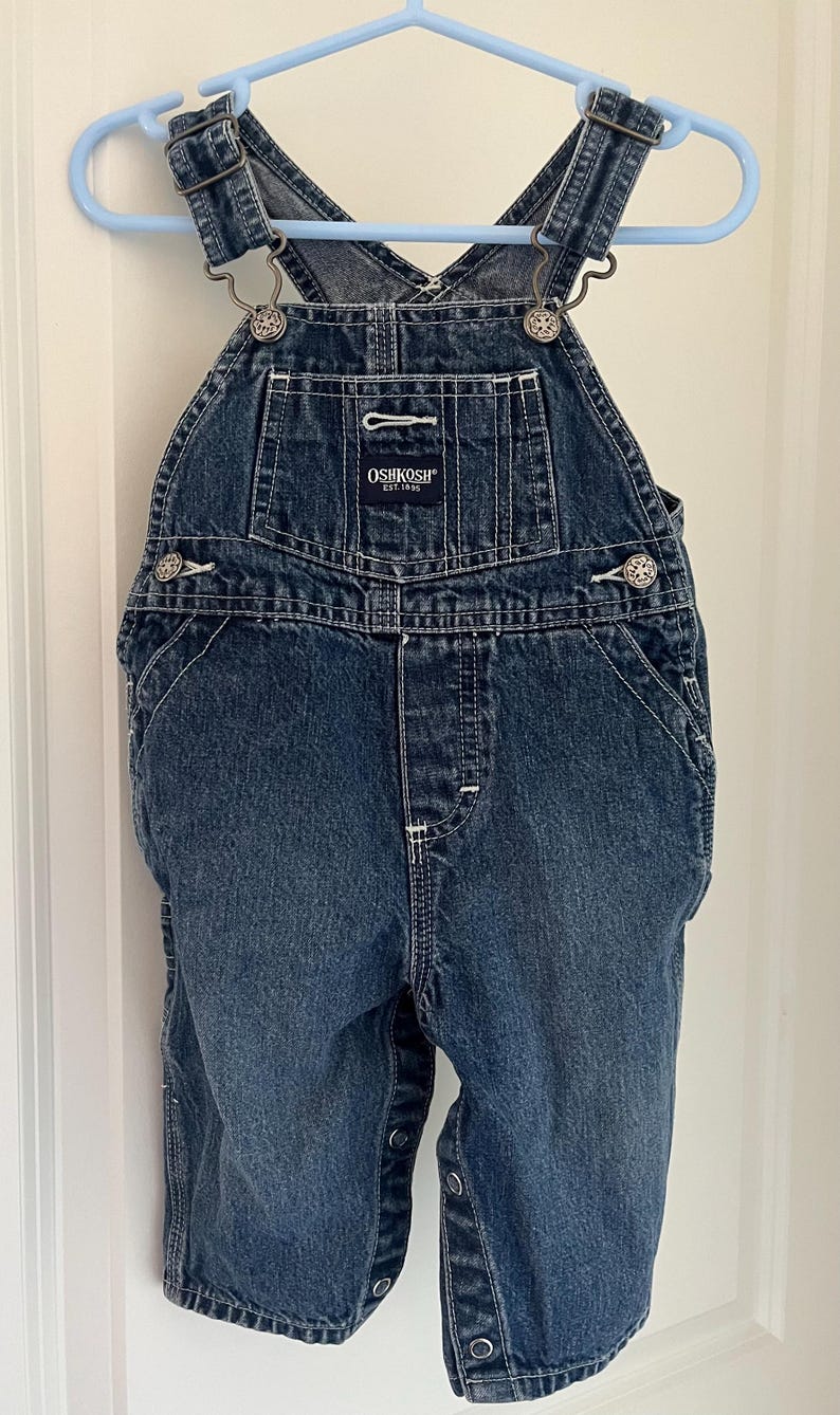 Puede incluir: Pantal&oacute;n vaquero azul para beb&eacute; o ni&ntilde;o peque&ntilde;o con tirantes ajustables y herrajes plateados. El pantal&oacute;n tiene el logotipo de OshKosh B'Gosh en la parte delantera con el texto "EST. 1895".